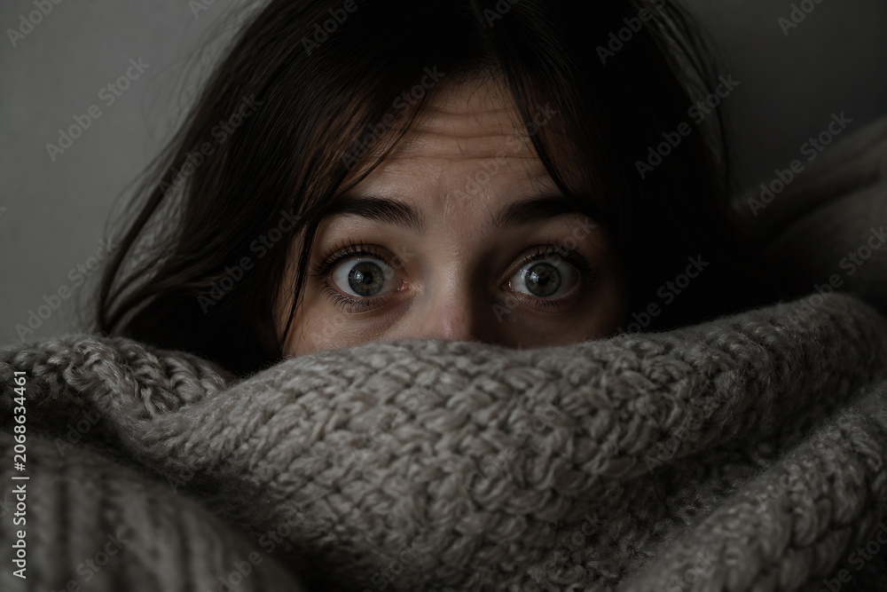 Fototapeta premium A person with wide, startled eyes peeking out from under a thick, knitted blanket in dim lighting.