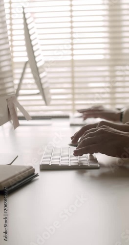 Close-up side view of a person's hands typing on a wireless keyboard at a minimalist office desk, with another person working on a computer in the blurred background near a window. vertical