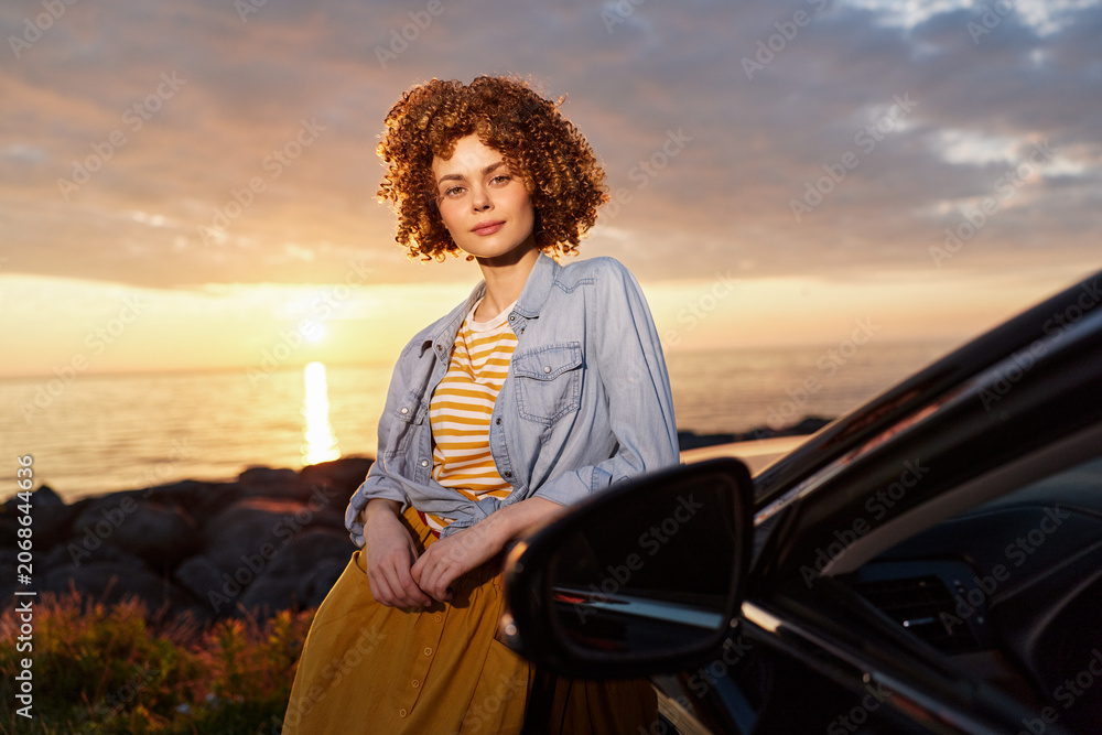 Fototapeta premium Young woman with curly hair leans on a car by a rocky shore at sunset, wearing a denim jacket over a striped top and orange skirt, radiating relaxed confidence