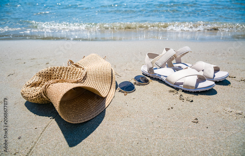 A straw hat, sunglasses, and white sandals are placed on sandy beach near the water. The scene captures a relaxed summer vibe.