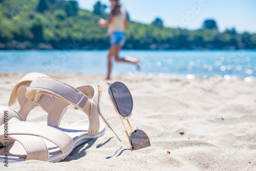 Sunglasses and sandals on a sandy beach. In the background, a young woman runs near the water. A bright sunny day with a clear blue sky.