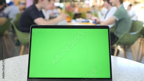 Laptop with green screen on cafe table. Blurred people sit in background. Focus remains on laptop screen. Ideal for digital mockups or presentations. Suitable for marketing or tech projects