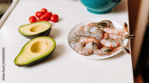 Fresh shrimp on white plate with avocado halves and cherry tomato on kitchen counter