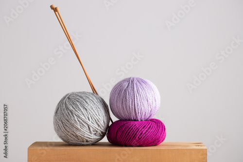 Three yarn balls in gray, lilac and fuchsia with knitting needles on light wooden base. Horizontal minimalist image with neutral background. Copy space. Concept of handmade craft and cozy lifestyle