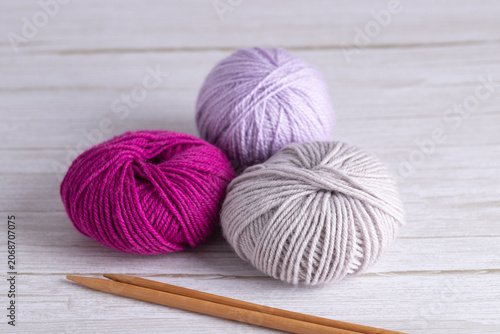 Three yarn balls in fuchsia, lilac and gray tones with wooden knitting needles on light gray wooden surface. Horizontal minimalist image with copy space