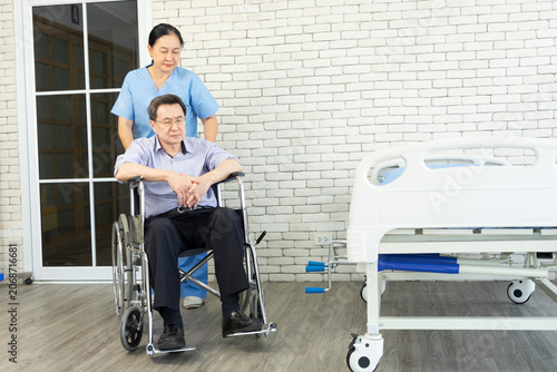 Smiling nurse assisting senior man to get up from bed. Caring nurse supporting patient while getting up from bed and move toward wheelchair at hospital. Helping elderly disabled asian man standing up.