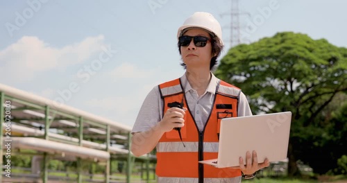 Asian adult female technician speaks through walkietalkie while holding laptop at oil refinery, managing emergency communication, pipeline safety inspection, compliance monitoring workflow report
