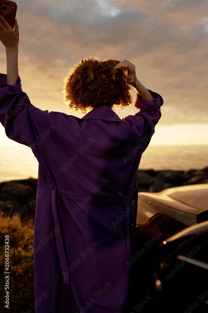 Fototapeta premium A woman wearing a purple robe stands with her back to the camera beside a car at sunset, raising her arms in a moment of reflection, freedom, and self care.