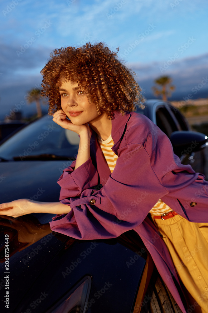 Obraz premium A stylish woman leans on a sleek car at dusk, wearing a purple jacket and casual outfit. She smiles softly, capturing a relaxed, confident lifestyle moment in the evening light.