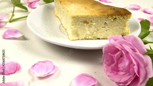 A white plate with a piece of homemade cottage cheese pie on it stands on a white table among roses and petals, side view