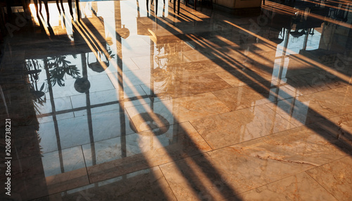 Long, dramatic shadows stretch across a highly polished, light-colored marble floor, reflecting interior elements and bright sunlight streaming through windows.