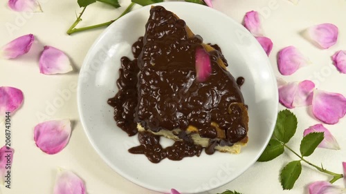 Edible rose petals fall on a piece of a cottage cheese pie with chocolate icing. White plate with a piece of a cottage cheese pie sits on a white table, among roses and petals, top view