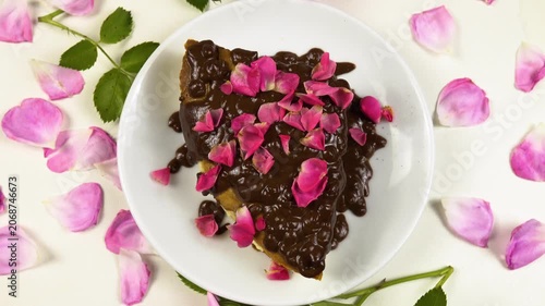 White plate with a piece of a cottage cheese pie with chocolate icing and edible rose petals sits on a white table, among roses and petals, top view