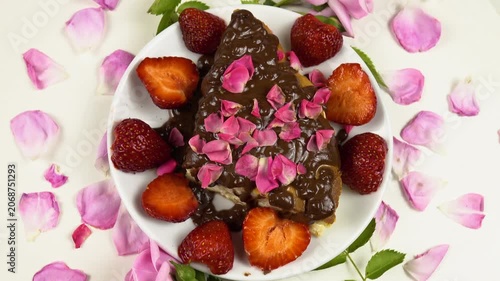 Plate with a piece of a cottage cheese pie with chocolate icing, rose petals and fresh strawberries sits on a white table, among roses and petals, top view
