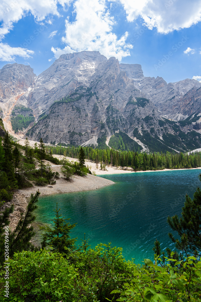 Obraz premium Turquoise water of Lake Braies with gravel shore surrounded by green pine forests and towering rocky mountain cliffs under a cloudy blue sky, scenic Pragser Wildsee landscape