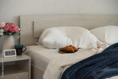 Croissants and coffee beside an unmade bed with a digital clock in a bright bedroom interior. Morning routine, hospitality and accommodation concept.