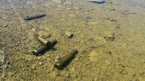 Several spent bullet casings lying at the bottom of a clear, shallow body of water with a muddy and rocky riverbed, suggesting a crime scene or evidence of a recent conflict or shooting