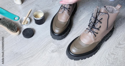 Woman hands wiping brown leather boots with grey microfiber cloth on floor. Female person buffing winter footwear using professional care products during domestic routine. Process of polishing leather