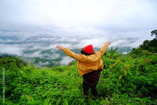 Asian young woman spread her arms smiling happy and wearing red hat and yellow jacket standing on peak of mountains and mist in the morning of Thailand 