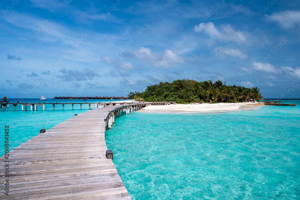 Fototapeta premium A wooden boardwalk curves over shallow turquoise water toward a palm island in the Maldives, with overwater villas on the horizon under bright midday sun.