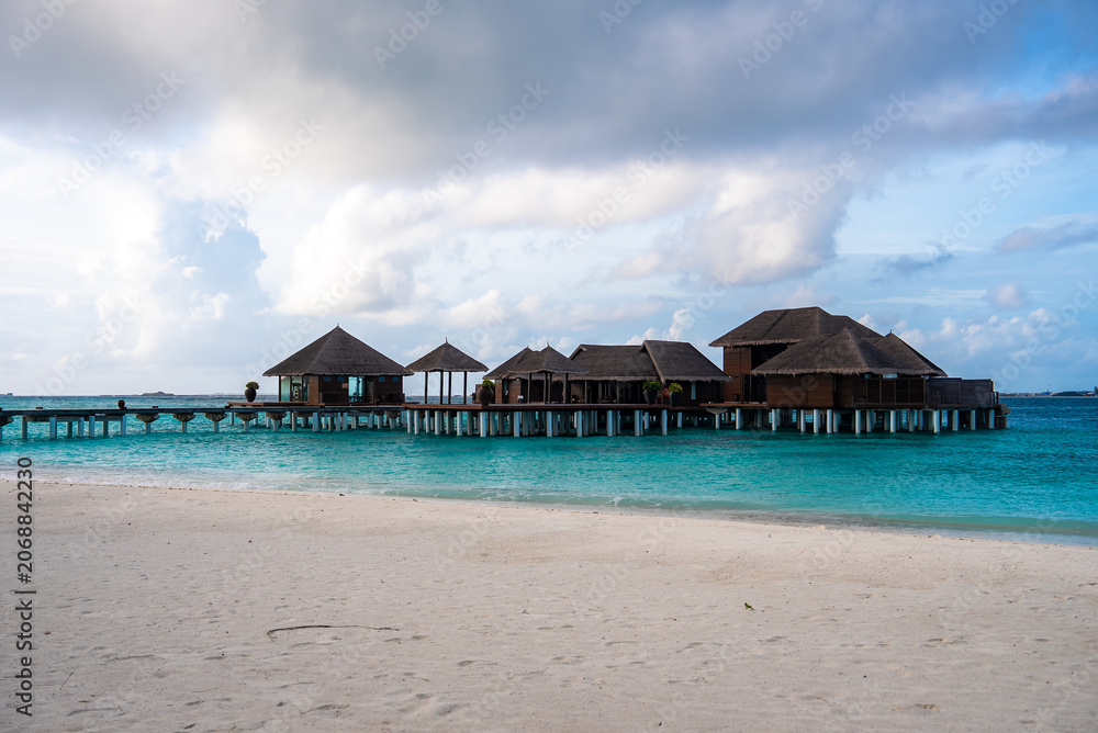 Fototapeta premium Thatched roof water villas on stilts connect by a long wooden boardwalk in the Maldives. Powdery white sand, calm seas, and cloud dappled skies frame the open horizon.