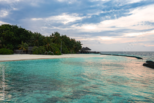Cuadro en lienzo A Maldives resort island shows a curving shore, powdery sand, dense palms, and a long wooden jetty to an overwater pavilion, with a reef breakwater under soft light