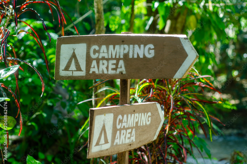 Fototapeta premium Brown wooden signs indicate camping area direction against a backdrop of vibrant green forest foliage for outdoor recreation planning