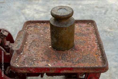 High angle close up of a single antique brass counterweight sitting on the rusty metal plate of a traditional red mechanical balance scale, conceptual business trade and weight measures backdrop views