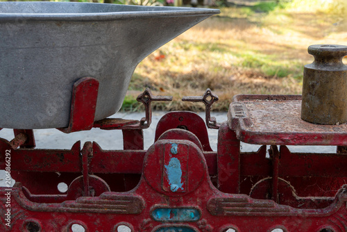 Close up views of a vintage traditional red iron mechanical balance scale with metal tray and heavy brass weight under natural outdoor morning daylight, rustic grocery market trade conceptual backdrop