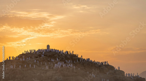 Sacred Mount Arafat sunrise captures pilgrims silhouettes against bright morning sky. Glowing clouds illuminate spiritual gathering on Mount Arafat.