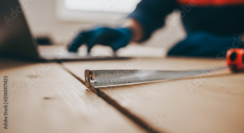 Metal tape measure on wooden planks with worker using laptop in background representing precision construction planning
