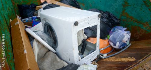 Discarded white washing machine inside a dumpster surrounded by bags of trash and cardboard boxes in an urban environment