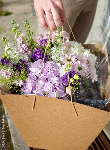 Hand holding a paper bag filled with colorful fresh flowers including purple, white, and pink varieties, with greenery visible, against a textured wall background