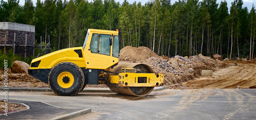 Yellow road roller compactor parked on a construction site with piles of earth and trees in the background, showcasing heavy machinery in action
