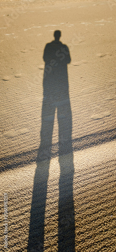 Shadow of a person standing on sandy beach during sunset, with long shadows cast on the textured sand surface creating a striking visual contrast