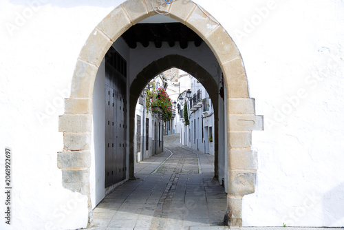 A view through the historic stone archway known as the Little Arch of Bread looking towards a charming cobblestone street lined with traditional white houses in Zafra, Spain.