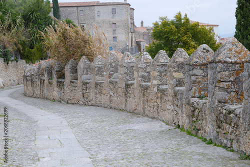 A curved historic cobblestone path running alongside the medieval stone defensive walls topped with pointed battlements in the old town of Trujillo, Caceres, Spain.