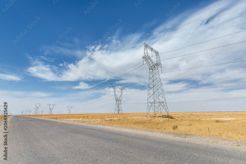Fototapeta premium Long Desert Road Parallel to High-Voltage Transmission Towers Under Blue Sky