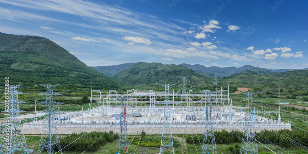 Fototapeta premium High-Voltage Substation Nestled in Green Mountain Valley Under Bright Blue Sky