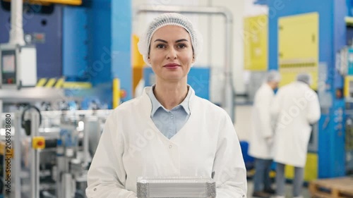 Portrait of Caucasian female factory manager wearing lab coat and hairnet proudly holding stack of aluminum containers. Standing in front of industrial machinery.