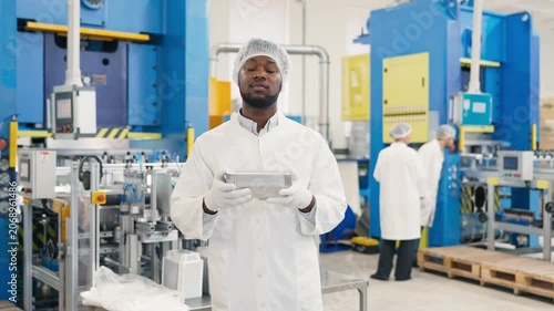African American male worker in protective uniform holds stack of aluminum containers. Standing in clean industrial factory environment with colleagues working in background.