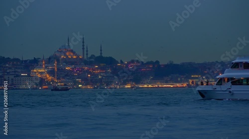 A beautiful twilight cityscape featuring a glowing mosque with minarets and urban lights across the water with a white boat cruising in the foreground in Istanbul