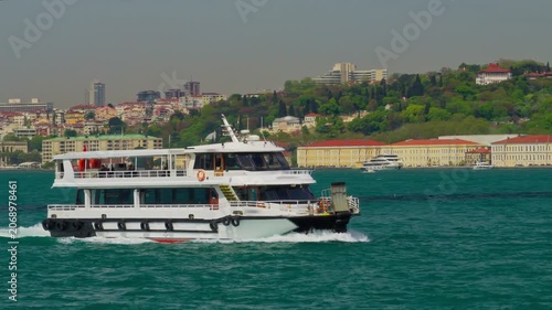 A white motorboat vessel moving through turquoise waters with a wake, featuring the scenic cityscape and urban architecture of Istanbul in the background under bright daylight