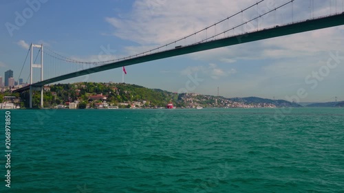 A wide shot of a massive suspension bridge spanning across the blue waters of the Bosphorus in Istanbul with green hills and urban coastline under a clear sky