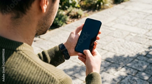 Young man wearing a green knitted sweater holds a modern black smartphone in his hands to check digital notifications while standing outside on a sunny afternoon during a bright day.