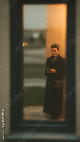 Smiling woman wearing a black coat using her smartphone reflected in a window glass pane