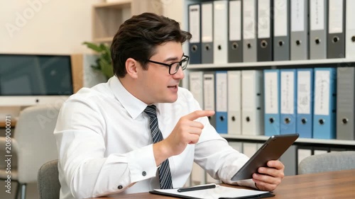 Young professional male employee wearing glasses and white shirt, engaged in a virtual meeting or video call with a tablet, seated at office desk surrounded by folders and documents.