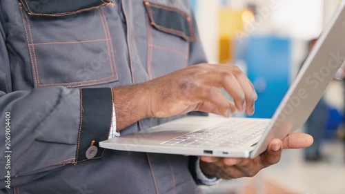 Close up of male hands typing on laptop keyboard entering production information. Wearing factory uniform. Worker using modern technology for data collection.