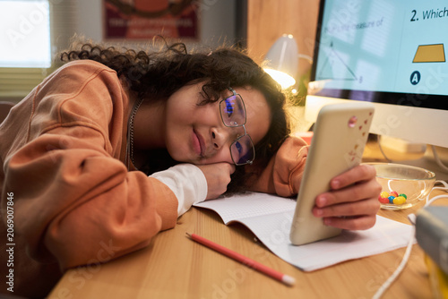 Obraz na płótnie Asian teenage girl resting on desk while holding smartphone during online study at home, showing screen distraction and digital learning fatigue