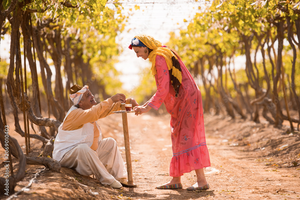 Fototapeta premium Authentic moment of Middle Eastern woman in vibrant traditional clothing serving tea to elderly man resting with walking stick on rural dirt path